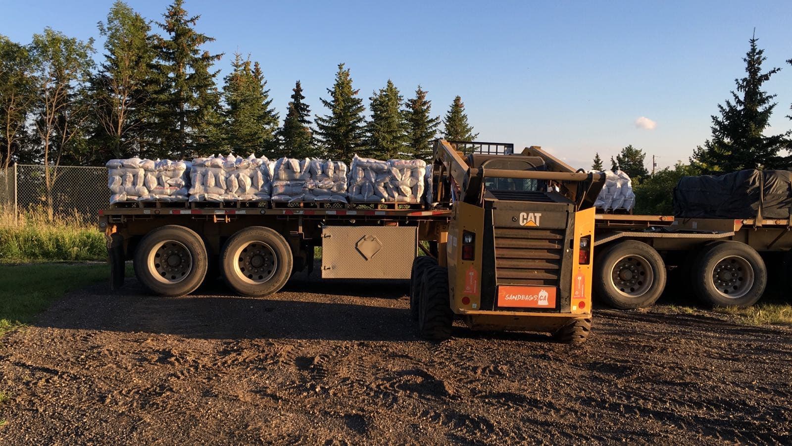 Flatbed trailer with pallets of sandbags and a skid steer at the yard